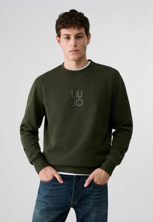Jeune homme portant un sweat-shirt vert foncé avec le logo "LIU JO" et un jean bleu, debout devant un fond gris clair uni.