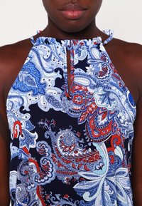 Haut à bretelles bleu marine à motif avec un design paisley bleu et rouge. Présente une petite ouverture en forme de clé et un col volanté. Fabriqué en tissu lisse.