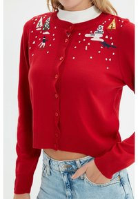 Cardigan rosso lavorato a maglia con maniche lunghe, chiusura con bottoni e motivi invernali ricamati tra cui alberi, uno sciatore e fiocchi di neve.