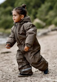 Bruine waterdichte skipak met capuchon en elastische manchetten, voorzien van een ritssluiting aan de voorkant en reflecterende accenten, gedragen door een kind buiten.