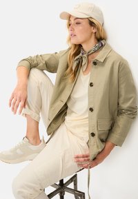 Lys olivengrøn jakke med brune knapper, creme top, creme bukser, lyse sneakers og en beige kasket. Bæres med et mønstret tørklæde.
