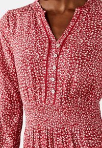 Rotes Kleid mit einem strukturierten Muster aus weißen Sprenkeln, mit durchgehender Knopfleiste, gerafftem Taillenbereich und volantiertem Ausschnitt.