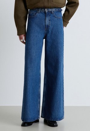 Persona che indossa jeans blu a vita alta e gamba larga con cintura nera, top marrone, scarpe nere e anello d'argento sulla mano sinistra.