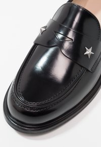 Mocassin en cuir noir poli avec des clous argentés en forme d'étoile et une découpe décorative sur la bride traversant le dessus.