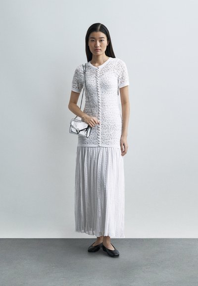 Γυναίκα με λευκή δαντελένια μπλούζα και πλισέ μακρύ φούστα κρατάει ασημένια τσάντα με αλυσίδα, στέκεται σε γκρι πάτωμα μπροστά σε απλό φωτεινό φόντο.