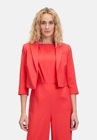 Blazer rosso su misura con maniche tre quarti e scollo tondo, indossato sopra una tuta coordinata. Tessuto morbido, dettagli minimi.