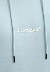 Sweat à capuche bleu clair en tissu doux, doté d'une poche kangourou et d'un cordon de serrage. Présente le texte blanc "JJ Studios" et le logo à l'avant.