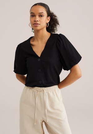 Frau mit zusammengebundenen lockigen Haaren, die goldene Creolen, ein schwarzes Oberteil mit Puffärmeln und Knöpfen sowie beige Hosen mit Kordelzug trägt, steht vor einem schlichten Hintergrund.