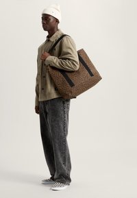 Man draagt beige jas, zwarte spijkerbroek, witte muts en geruite sneakers, met een grote bruine tas met luipaardprint over de schouder.