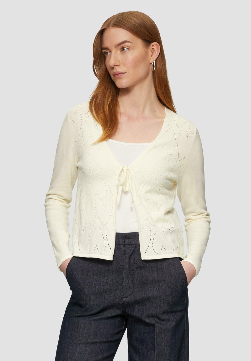 Gilet crème avec un motif en forme de cœur, texture tricotée, manches longues et fermeture à nouer à l'avant. Associé à un haut blanc et un pantalon sombre.
