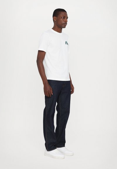 Camiseta de algodón blanca con un gráfico verde en el pecho, combinada con pantalones de mezclilla oscura y zapatillas blancas, diseño y ajuste minimalista.