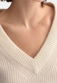 V-Ausschnitt Pullover in Cremefarbe, mit strukturiertem Rippenmuster und einem glatten, weichen Strick. Detail von Halsausschnitt und Schulter sichtbar.