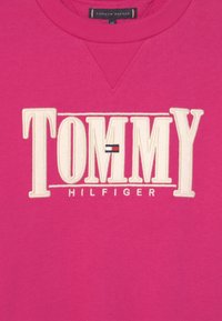 Sudadera rosa con un logotipo texturizado en blanco bordado de "TOMMY" y "HILFIGER" debajo, presentando un pequeño acento de bandera roja, blanca y azul.