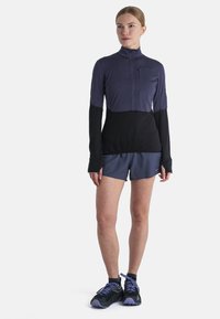 Langärmliges Sportoberteil in Purpur und Schwarz mit einem Reißverschluss am Hals. Kombiniert mit dunkelblauen Shorts und schwarzen Sportschuhen mit lila Akzenten.