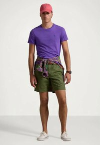 Άνδρας που φοράει μια μοβ μπλούζα, πράσινα σορτς, ροζ καπέλο, λευκά αθλητικά παπούτσια, ρολόι χειρός και καρώ πουκάμισο δεμένο γύρω από τη μέση.