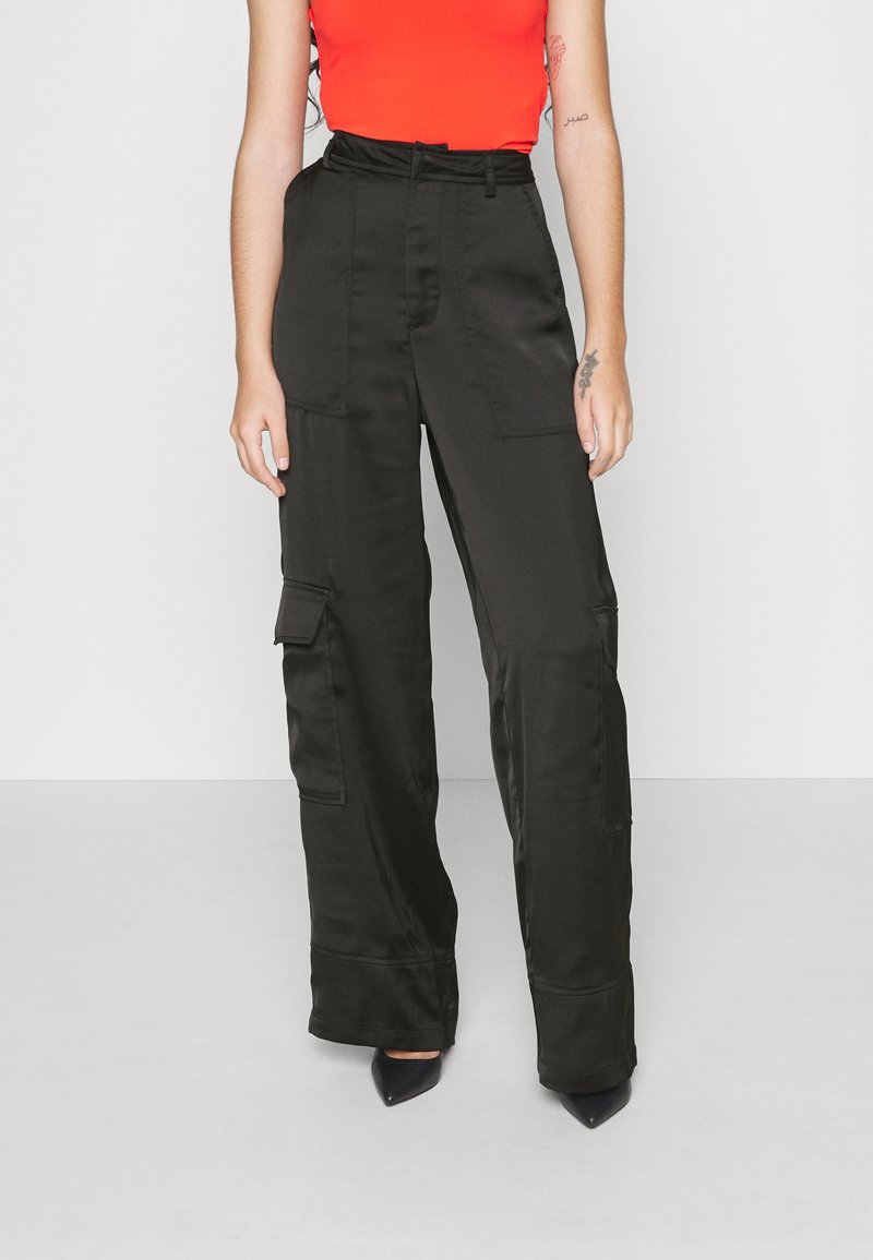 Personne portant un pantalon cargo noir taille haute à jambes larges, un top rouge sans manches et des chaussures noires à bout pointu sur fond blanc uni.