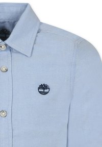 Chemise boutonnée bleu clair avec un col pointu, des boutons argentés et un logo de arbre noir brodé sur la poitrine gauche.