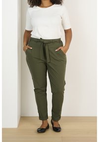 Pantaloni aderenti verde oliva con cintura anteriore a fettuccia, tasche laterali e polsini arrotolati, abbinati a una maglietta bianca a maniche corte e ballerine nere.