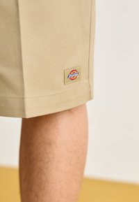 Tan shorts with a straight cut, featuring a visible "Dickies" label patch with a red, yellow, and blue logo near the hem.