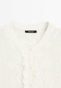 Cardigan blanc en maille avec motifs floraux, bordure froncée le long de la patte de boutons et col rond. L'étiquette indique « Massimo Dutti ».