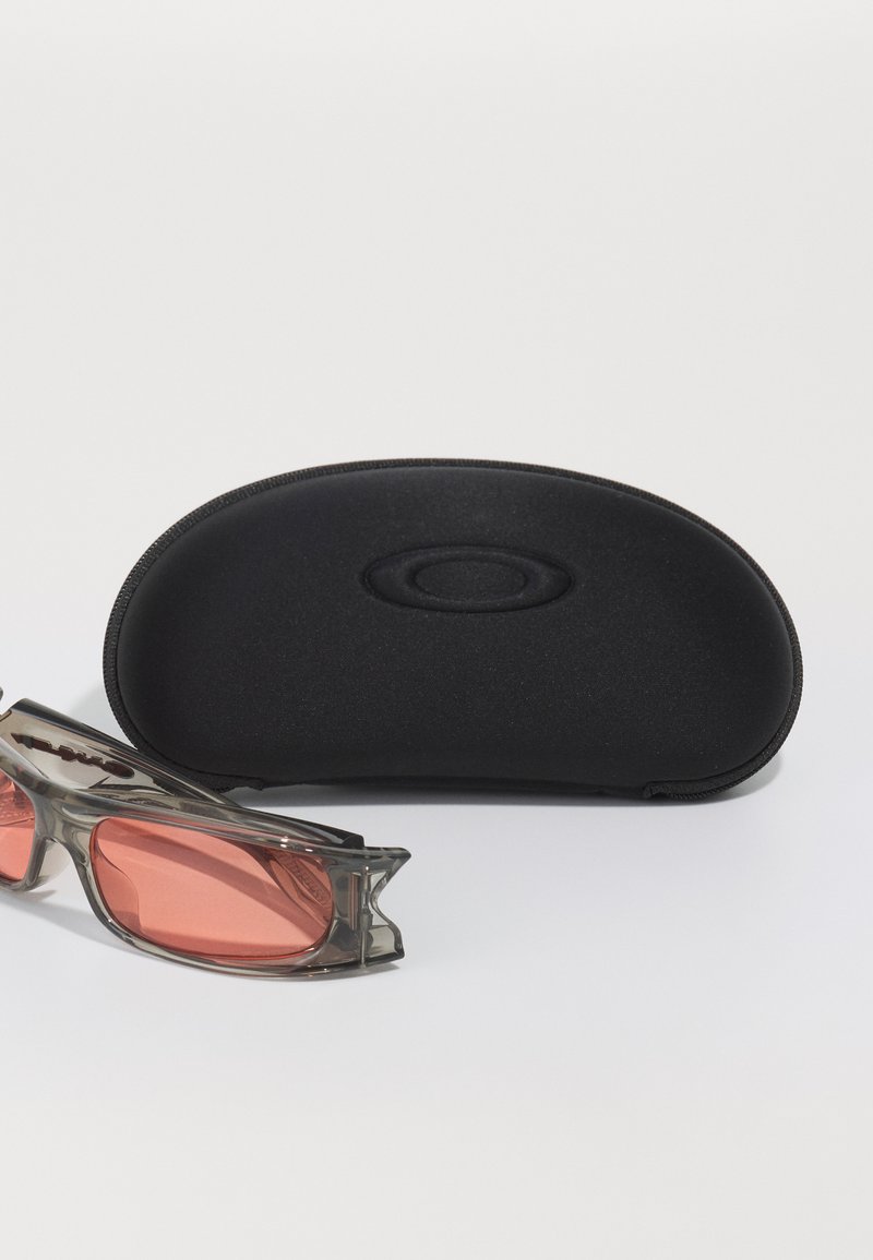 Occhiali da sole con montatura trasparente e lenti rosa accanto a un astuccio rigido protettivo black con il marchio Oakley su una superficie bianca.