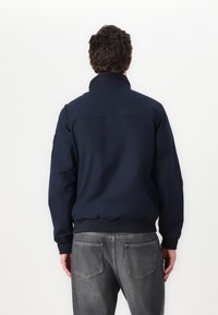 Giacca blu navy con colletto alto e polsini elastici, caratterizzata da una trama liscia e opaca. Indossata con jeans grigi, vista da dietro.