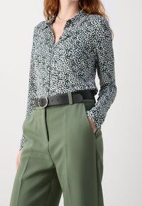 Groen gemustreerd overhemd met witte spikkels, gecombineerd met hooggesneden, geplooide olijfgroene broek en een zwarte riem met een ronde gesp.