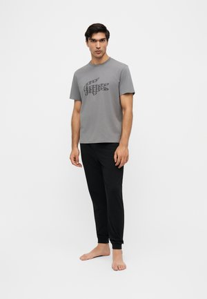 Mann steht barfuß, trägt ein graues T-Shirt mit einem geometrischen Krokodil-Design und schwarze Jogginghose vor einem einfarbigen weißen Hintergrund.