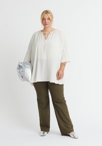 Weißes lockeres Hemd mit V-Ausschnitt und Bindedetail, kombiniert mit olivgrünen weiten Hosen. Das Model hält eine silberne Tasche und trägt metallische Schuhe.