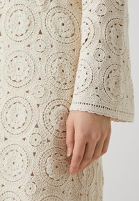 Robe en crochet ornée de motifs circulaires complexes en beige clair, avec une finition texturée et un design à manches évasées.