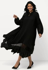 Vestido negro plisado con dobladillo con volantes, mangas largas y escote en forma de ojo de cerradura, combinado con tacones negros de punta contra un fondo neutro.