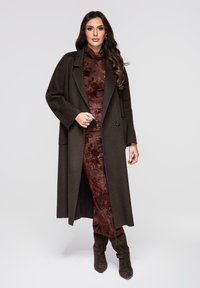 Abrigo largo de mezcla de lana marrón con solapas anchas sobre un vestido con patrón bronceado. Botas altas hasta la rodilla en marrón oscuro completan el conjunto.