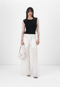 Black ribbed sleeveless top with gold accents, paired with white wide-leg trousers featuring patch pockets. Model holds a small white handbag.