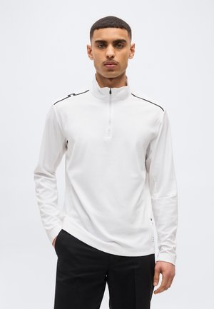 Mladý muž s krátkymi vlasmi, oblečený v bielom tričku s dlhým rukávom a štvrtinovým zipsom a čiernych nohaviciach, stojaci s jednou rukou v vrecku.