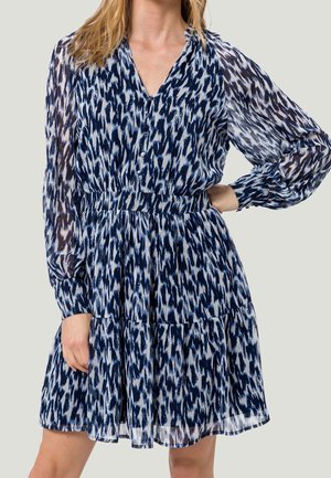 Frau trägt ein blau-weiß gemustertes, langärmeliges Kleid mit Knöpfen vorne und einem stufigen Rock.