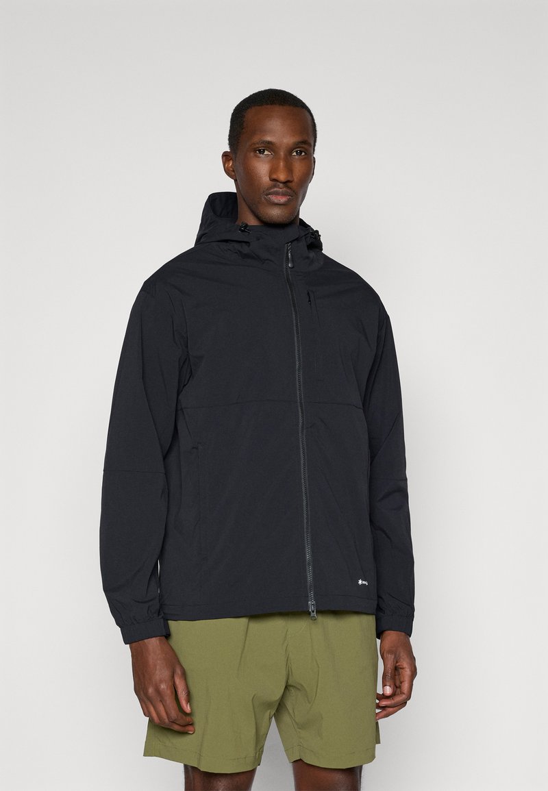 Veste légère noire avec fermeture éclair à l'avant, capuche ajustable et poignets élastiques, associée à un short vert olive.