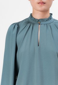 Kvinne iført en turkise bluse med lange puffede ermer, rynket høy hals og frontspalte sikret med en liten gullknapp.