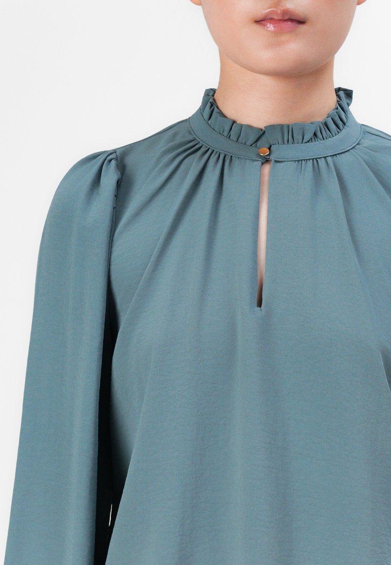 Vrouw in een teal blouse met lange, opgezette mouwen, gerimpelde hoge halslijn en een voorkant met split dat is beveiligd met een kleine gouden knop.