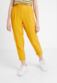 Gula avsmalnande byxor med hög midja, plisserad design och uppvikta benslut. Bärs med en randig topp och vita sneakers.