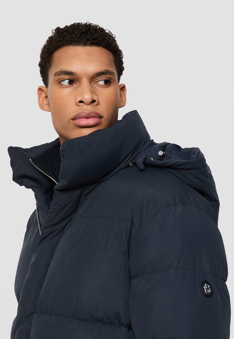 Veste matelassée bleu marine avec un col haut, fermeture éclair à l'avant et capuche avec cordon de serrage. Possède une texture matelassée et un logo en patch sur le côté.