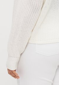 Weißer Strickpullover mit strukturiertem Muster, gerippten Bündchen und figurnahen Design, kombiniert mit weißen Jeans, die am unteren Rand des Bildes sichtbar sind.