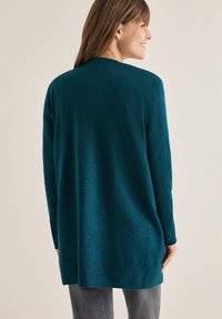 Tyrkysový dlouhý kardigan s pletenou texturou, kulatým výstřihem a žebrovanými manžetami. Spodek sahá pod boky, což zajišťuje volný střih.