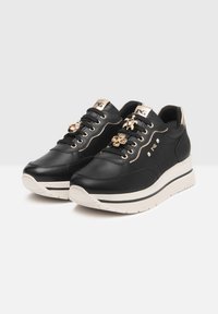 Czarne platformowe sneakersy z beżowymi podeszwami, złotymi oczkami i dekoracyjnymi złotymi zawieszkami w kształcie żółwi i kwiatów na czarnych sznurówkach.