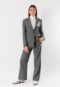 Completo grigio a righe con un blazer su misura che presenta una toppa testurizzata. Abbinato a pantaloni larghi e sneaker bianche, su uno sfondo semplice.