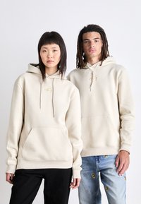 Beige hoodies med dragsnören, framfickor och broderad logotyp. Den manliga modellen bär denimjeans med detaljer; den kvinnliga modellen har svarta byxor.