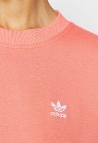 Korálově růžová žebrovaná mikina s bílým logem Adidas trefoil na levé straně hrudi. Má kulatý výstřih a měkkou texturu.