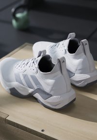 Coppia di scarpe sportive Adidas bianche e grigie con linguette sul tallone, posate su una superficie in legno con un kettlebell sfocato sullo sfondo.