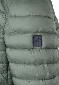 Groene pufferjack met horizontale stiksels, met een gladde textuur. Inclusief een rechthoekig navy stoffen label op de linkermouw.