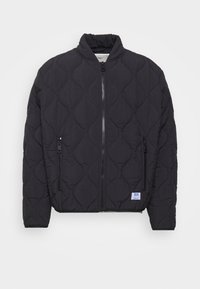 Musta tikk quiltattu bomber-takki, jossa on korkea kaulus, vetoketjullinen etuosa ja kaksi sivutaskua. Alhaalla vasemmalla sivulla on merkkitikkaukset.