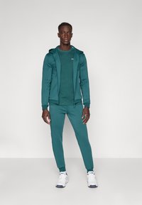 Tenue de sport teal composée d'un hoodie zippé, d'un t-shirt et d'un pantalon de jogging. Comprend des poignets et des poches latérales. Associée à des baskets blanches.
